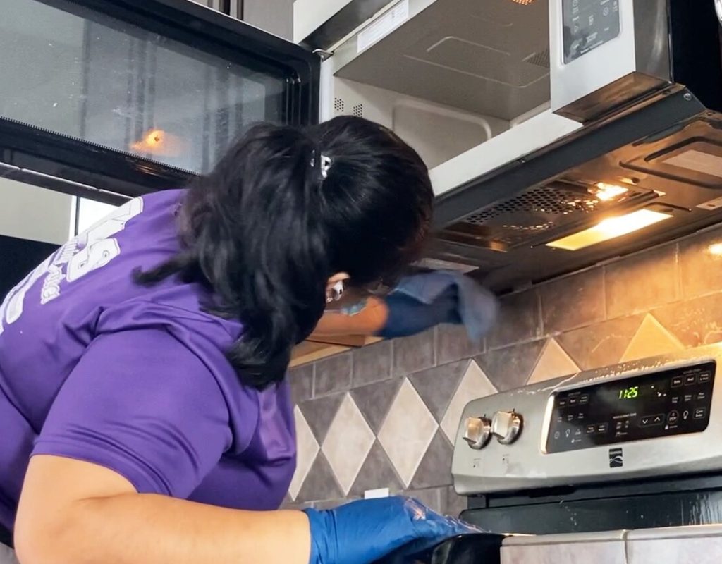 Reliable weekly cleaning in Charlestown, IN with a professional cleaner wiping and sanitizing a residential microwave, stovetop, and kitchen backsplash.
