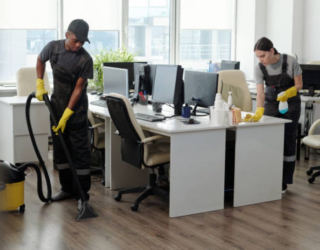 Young male and female workers of service company using detergent and vacuum cleaner for cleaning contemporary openspace office